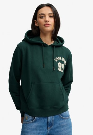 Młoda kobieta o prostych ciemnych włosach, ubrana w ciemnozieloną bluzę z kapturem z napisem "SUPERDRY 89" i niebieskie jeansy na jednolitym tle.