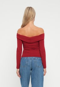 Top rojo sin mangas con un hombro al descubierto y mangas largas, que presenta una textura fruncida en la parte posterior, combinado con unos vaqueros de mezclilla azul de cintura alta.