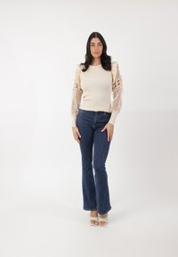 Maglione beige a coste con maniche lunghe in pizzo trasparente, abbinato a jeans a palazzo blu scuro e sandali bianchi con cinturino. Sfondo bianco semplice.