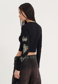 Zwart cropped long-sleeve top van textuur stof. Voorzien van grafische prints van een kroon en decoratieve motieven op de mouwen. Gecombineerd met donkere broeken.