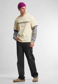 T-shirt couleur crème avec imprimé marron superposé sur une chemise à manches longues grise, associé à un pantalon en denim foncé et des sabots beiges. Bonnet en tricot violet.