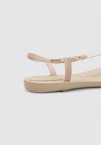 Sandalia de plástico beige con suela plana. Presenta un diseño de tiras delgadas, acentos metálicos y hebilla ajustable. Plantilla texturizada.