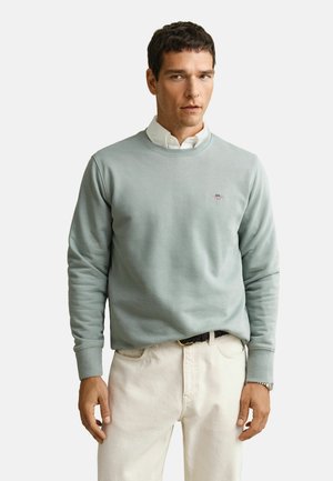 Mann, der ein hellblaues Sweatshirt mit Rundhalsausschnitt über einem weißen Hemd mit Kragen trägt, kombiniert mit cremefarbenen Hosen und einem schwarzen Gürtel, steht vor einem schlichten Hintergrund.
