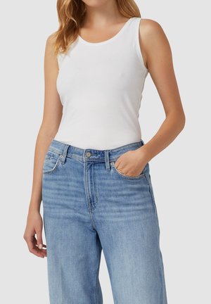 Femme portant un haut blanc sans manches ajusté, rentré dans un jean en denim bleu clair taille haute, avec une main dans la poche.