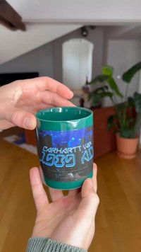 Grønn keramikkrus med svart grafisk design og blå tekst som sier "Carhartt WIP 1989 A.D." Hånd som holder koppen mot en uskarp bakgrunn.