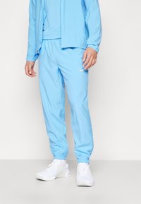 Lichtblauwe sportjas met bijpassende broek, voorzien van elastische manchetten en zoom, gedragen met witte sneakers. Eenvoudig ontwerp, gladde textuur.