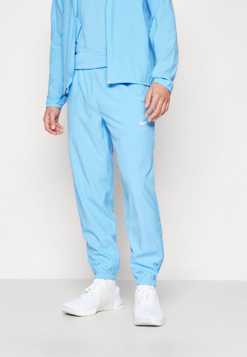 Lichtblauwe sportjas met bijpassende broek, voorzien van elastische manchetten en zoom, gedragen met witte sneakers. Eenvoudig ontwerp, gladde textuur.