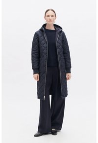 Abrigo largo acolchado en navy con capucha, cierre de cremallera y bolsillos laterales. Lleva puesto sobre un suéter oscuro y pantalones de pierna ancha. Textura suave y acolchada.