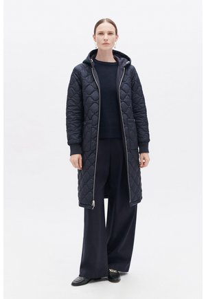 Quilted navyblauer langer Mantel mit Kapuze, Reißverschluss und seitlichen Taschen. Getragen über einem dunklen Pullover und weit geschnittenen Hosen. Glatte, gepolsterte Textur.
