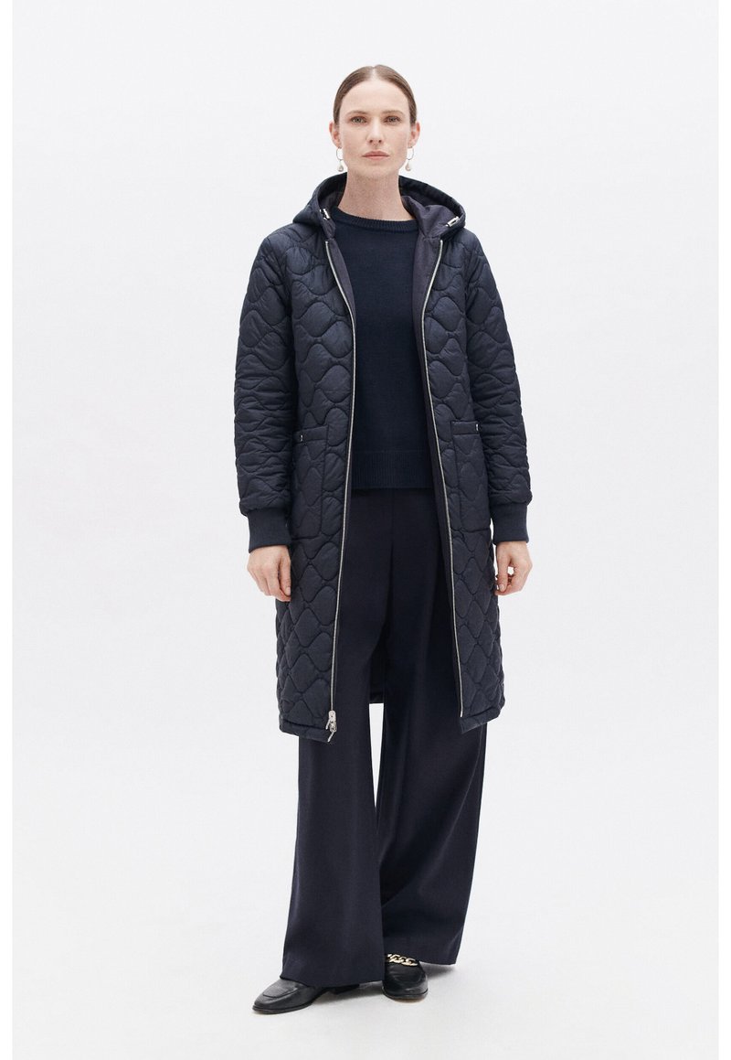 Abrigo largo acolchado en navy con capucha, cierre de cremallera y bolsillos laterales. Lleva puesto sobre un suéter oscuro y pantalones de pierna ancha. Textura suave y acolchada.