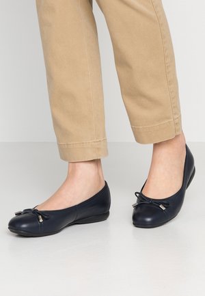 Navyblauwe leren ballerina's met een ronde neus, decoratieve strik en zilveren accenten. G gedragen met kaki tapered broek tegen een lichte achtergrond.