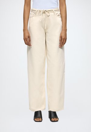 Άτομο που φοράει μπεζ παντελόνες με ψηλή μέση και φαρδιά ποδόγυρα με κορδόνι στη μέση, συνδυασμένες με μαύρα πέδιλα μέσης φτέρνας με ανοιχτά δάχτυλα.