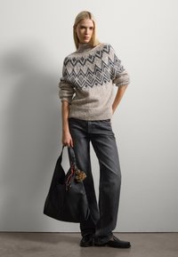 Maglione beige voluminoso con pattern geometrici scuri, abbinato a pantaloni grigi a gamba larga. La modella tiene una borsa a tracolla in pelle nera con un pompon.