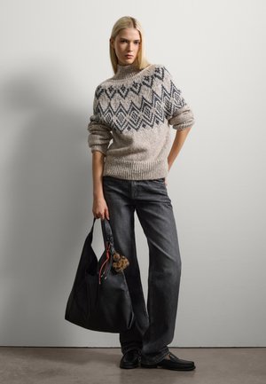 Chunky beige trui met donkere geometrische patronen, gecombineerd met grijze wijde jeans. Model houdt een zwarte leren tote bag met een kwast vast.