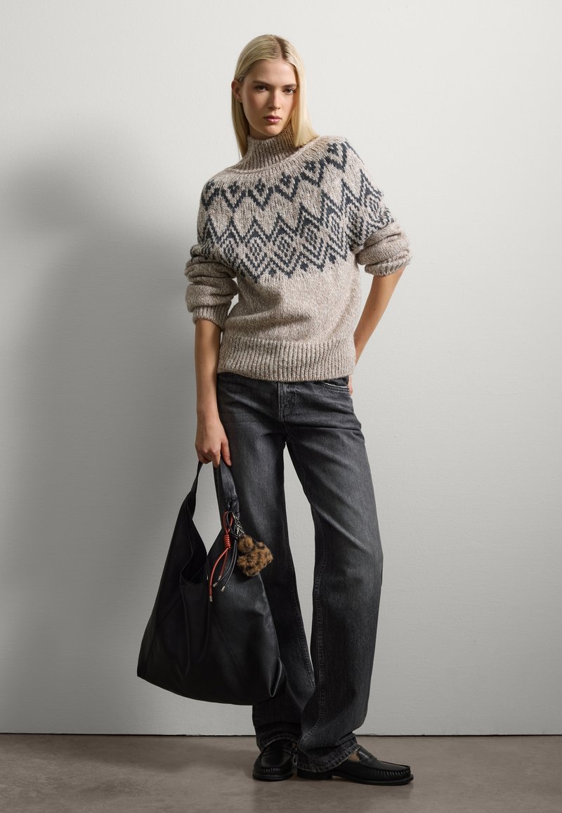 Maglione beige voluminoso con pattern geometrici scuri, abbinato a pantaloni grigi a gamba larga. La modella tiene una borsa a tracolla in pelle nera con un pompon.