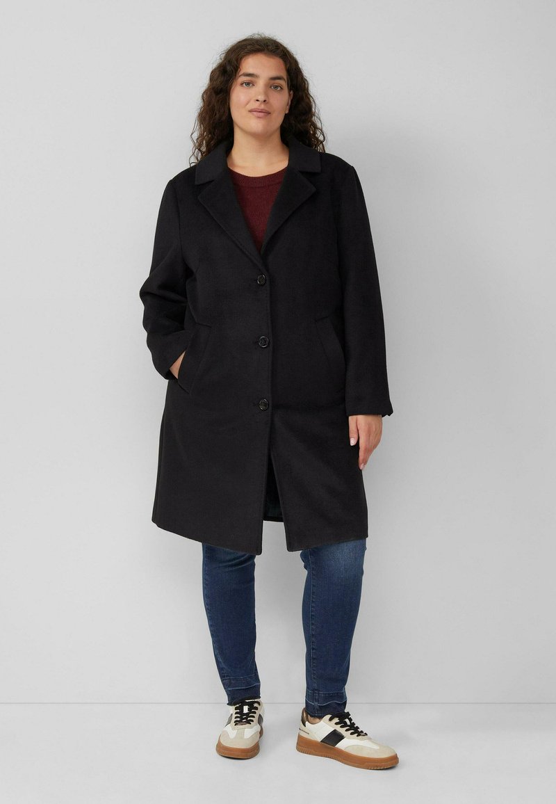 Zwarte wollen jas met een notch-kraag, knoopsluiting en zijzakken. gedragen over een bordeauxrode trui en gecombineerd met blauwe jeans en sneakers.