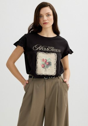 Joven mujer con cabello largo y oscuro lleva una camiseta negra "Warm Memories" con gráfico de fresa y pantalones beige de cintura alta, manos en los bolsillos.