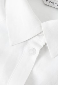 Camicia bianca in lino con colletto a punta, dotata di due bottoni e un tessuto texturizzato. Etichetta visibile con il logo del brand.