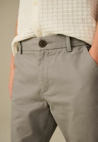 Un pantalon en coton gris présente une texture lisse, une fermeture à bouton et des poches latérales pour plus de praticité. La taille est ajustée pour un bon maintien.