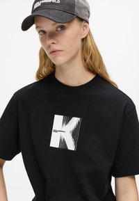 Juodas medvilninis marškinėliai su baltu grafiniu piešiniu, kuriame pavaizduota "K" ir linijos, apvaliu kaklu ir trumpomis rankovėmis. Dėvimi su pilku kepurėle.