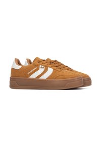 Zapatillas de ante color beige con un talón de cuero blanco y tres franjas blancas en el costado. Presentan una suela de goma y una lengua acolchada para mayor comodidad.