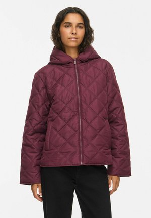 Donna che indossa una giacca trapuntata con cappuccio color borgogna con cerniera frontale e pantaloni neri su sfondo semplice.