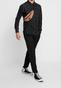 Bruine ribstof schoudertas gedragen over een zwart denim overhemd, in combinatie met zwarte broek en zwarte hoge sneakers met witte accenten.