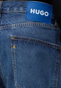 Μπλε τζιν από denim με ορατά ραφές στις πίσω τσέπες και μπλε ορθογώνιο πλακίδιο με την επιγραφή HUGO στη μέση.