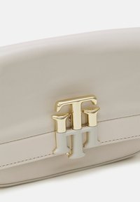 Beige leren tas met een gebogen vorm, voorzien van een opvallende goudkleurige logo-sluiting en een soepele textuur. Grijze accenten complementeren het ontwerp.