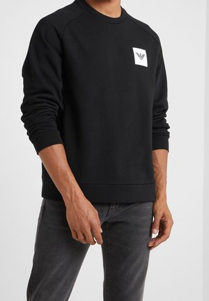 Mand iført en sort langærmet sweatshirt med et lille hvidt logo på venstre bryst, kombineret med mørkegrå jeans.