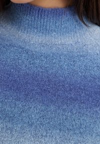 Blå stickad tröja med en ombré-gradient från mörka till ljusa nyanser, med en texturerad, mjuk kvalitet och hög krage.