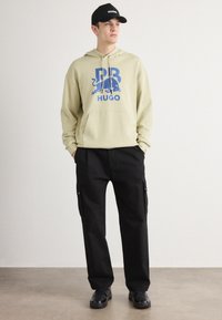 Felpa color beige chiaro con logo grafico blu, tasca frontale; abbinata a pantaloni cargo neri con tasche laterali e scarpe da ginnastica nere.