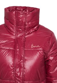 Doudoune bordeaux avec un col montant, un design matelassé, une texture brillante et un logo brodé sur la poitrine.