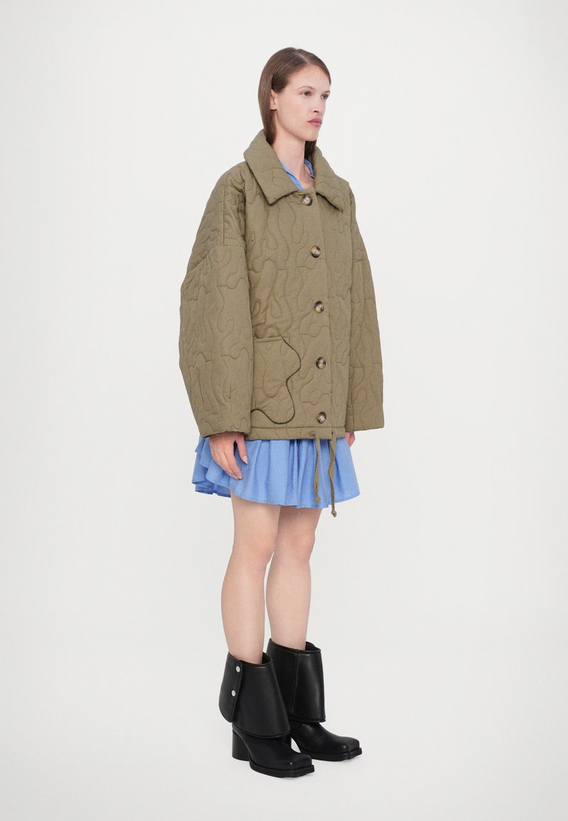 Veste matelassée vert olive avec grand col, boutons à l'avant et poches latérales, portée avec une robe bleu clair et des bottines noires.