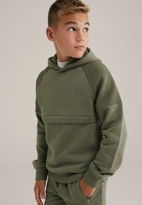 Zöld kapucnis pulóver texturált ujjal, elöl logóval és állítható kapucnival. Puha anyagú, bordázott mandzsettákkal, hozzáillő alsóval párosítva.