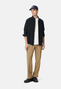 Camicia azzurra con bottoni sopra una maglietta bianca, pantaloni cargo beige, scarpe nere e un cappellino blu con dettagli testuali.
