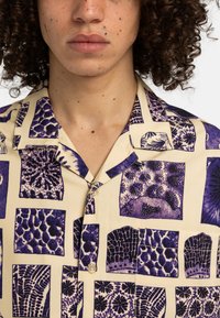 Short-sleeve shirt in cream with purple botanical print, featuring various patterns and button closure, made from lightweight fabric.