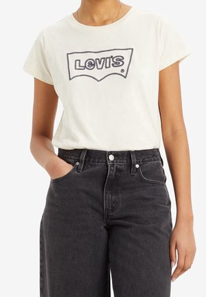 Persoon draagt een wit Levi's T-shirt met zwart gestikt logo en een zwarte high-waisted spijkerbroek, met één hand in de zak.