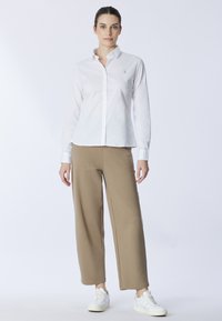 Camicia bianca con colletto e logo ricamato, abbinata a pantaloni beige a gamba larga e sneaker bianche, su uno sfondo neutro.
