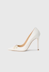 Λευκή γόβα από πατενταρισμένο δέρμα με ψηλό τακούνι και μυτερή μύτη, διακοσμημένη με χρυσό λογότυπο στο μπροστινό μέρος και κομψή, λεπτή σιλουέτα.