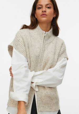 Beige gerippte Strickweste mit hohem Kragen und Reißverschluss, getragen über einem weißen Hemd mit Knöpfen. Streifen in unterschiedlichen Schattierungen verleihen Struktur.