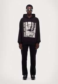 Felpa nera con stampa grafica e testo bianco, abbinata a pantaloni neri e scarpe sportive nere. Il modello indossa occhiali da sole bianchi.