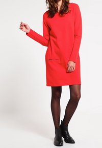 Rode jurk met lange mouwen en een rechte snit, voorzien van ritszakken en een ronde halslijn. Zwarte enkellaarsjes maken de outfit compleet.