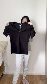 Personne tenant un t-shirt noir col V devant un mur blanc, portant un pantalon de jogging gris et des chaussons roses dans une chambre.