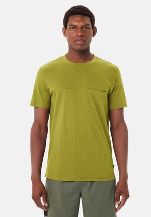 Junger Mann, der ein olivgrünes kurzärmeliges T-Shirt und dazu passende grüne Hosen trägt, vor einem schlichten hellen Hintergrund steht.