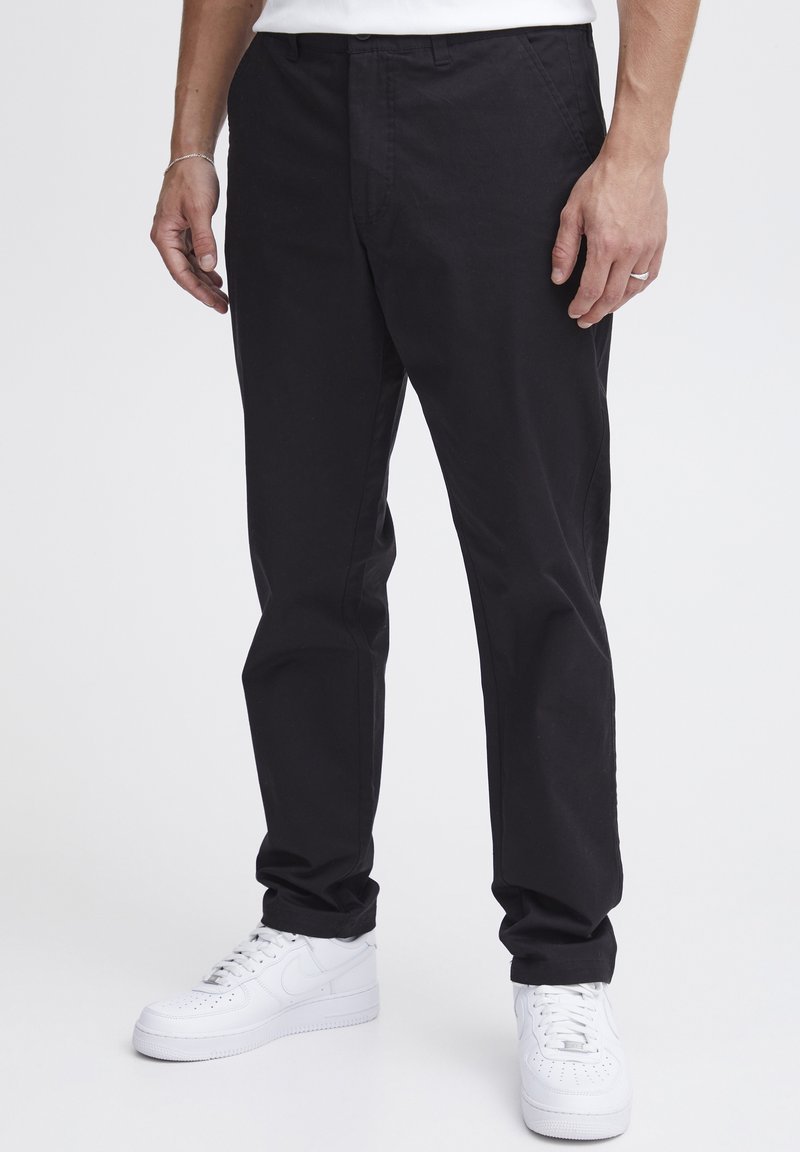 Pantaloni negri drepți, cu o textură netedă, având buzunare laterale și închidere cu nasture. Purtați cu adidași albi.
