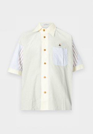 Jonge man die een gestreept kort mouw overhemd met knopen draagt, beige broek en gele loafers, staand tegen een effen lichte achtergrond.
