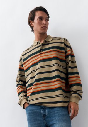 Νέος άνδρας που φοράει ένα μπεζ πόλο με μακριά μανίκια και οριζόντιες πράσινες, πορτοκαλί και μαύρες ρίγες, συνδυασμένο με μπλε τζιν, στέκεται μπροστά σε έναν λευκό τοίχο.