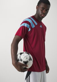 Rotes Sportshirt mit blauen Akzenten und drei Streifen auf der Schulter, mit einem runden Halsausschnitt. Das Model hält einen schwarz-weißen Fußball.