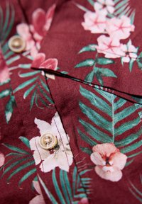 Bordeaux stof met een bloemen- en bladerenpatroon. Bevat crèmekleurige knopen en een gestructureerde weving. Close-up van de kraag en knoopdetails.